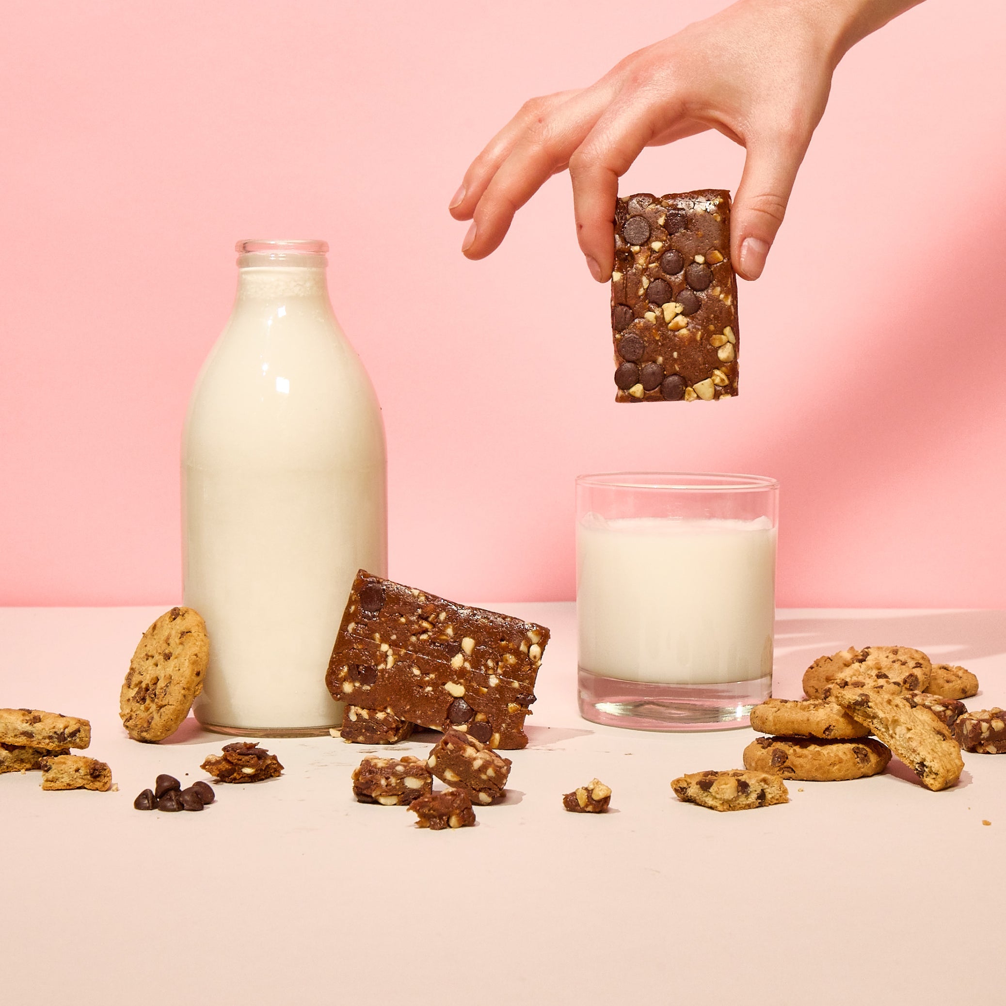 Hand holding Lucid's Cookie Dough Snack Bar, dipping into a glass of milk, alongside a milk bottle and surrounded by chocolate chip cookies.